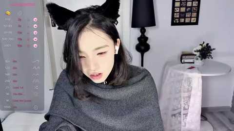 Media: Video of an Asian woman with light skin, wearing a gray blanket and black cat ears, sitting in a modern, white room with a black lamp and framed art on the wall.