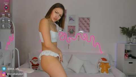 Media: A video of a young woman with fair skin, wearing white lingerie, posing seductively on a bed with a pink neon sign and holiday decor.