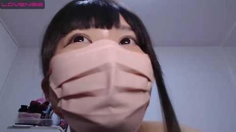 Media: Video of an Asian woman with straight black hair and bangs wearing a white face mask, looking up with wide eyes, in a plain white room.