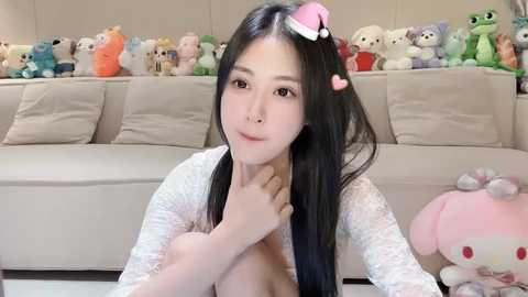 Media: A video of an Asian woman with long black hair, wearing a Santa hat, sitting topless on a beige sofa. She holds her breast. Background features stuffed animals.