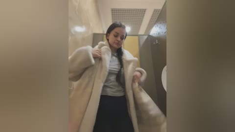 Media: A video shows a young woman with dark hair, wearing a beige fur coat, a gray sweater, and black pants, adjusting the coat in a narrow, beige-tiled hallway with fluorescent lighting.