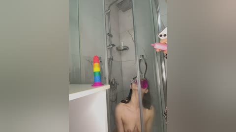 Media: Video of a naked woman with long black hair, wearing a pink face mask, in a modern, white-tiled shower, featuring colorful sex toys and a smartphone.