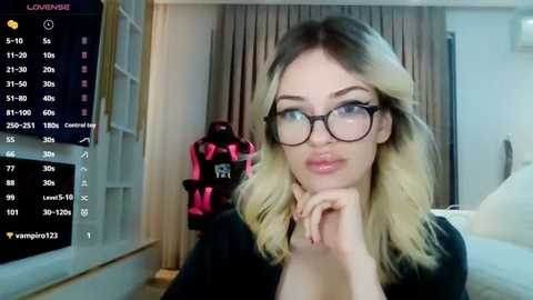 Media: Video of a blonde woman with glasses, resting her chin on her hand, in a modern bedroom with a gaming setup in the background.