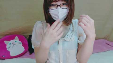 Media: Video of an Asian woman with glasses and a white mask, wearing a light blue shirt, sitting on a bed with pink and white pillows and a plush cat.