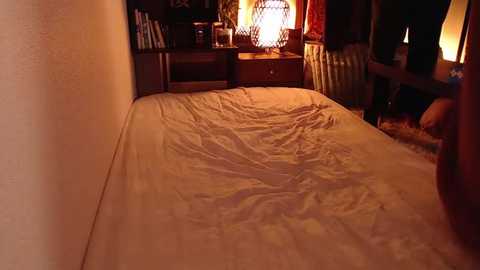 Media: Video of a dimly lit bedroom with a neatly made bed in the foreground, a wooden nightstand with a lamp, and a chair with a cushion in the background.