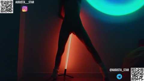 Media: A dimly lit video of a person in a tight, dark bodysuit standing in front of a glowing red lamp with a circular blue light in the background. The person's silhouette is visible, and the room has a minimalist, modern feel.