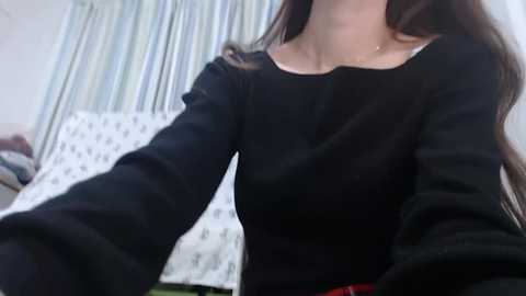 Media: Video of a woman with long brown hair, wearing a black long-sleeve shirt, standing in a bedroom with white bed sheets and blue striped curtains.