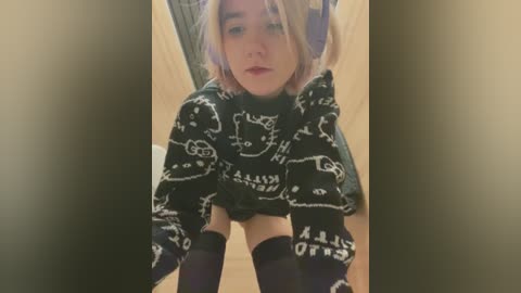 Media: Video of a pale-skinned person with shoulder-length blonde hair, wearing a black, cartoon-print sweater, black thigh-high socks, and sitting on a wooden bench. Background is blurry, indoors.