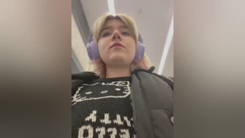 Media: A video of a young woman with light skin and shoulder-length blonde hair, wearing large purple headphones, black sweater with white patterns, and a gray puffy jacket, taken from a low angle, indoors with fluorescent lighting.