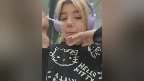 Media: A video of a young blonde woman with headphones, wearing a black sweater with a Hello Kitty design, holding a spoon while eating, with blurred people in the background.