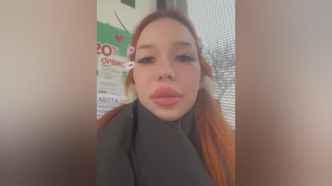 Media: Video of a young woman with fair skin, red hair, and full lips, wearing a black top, standing in a room with a poster and a mesh window.