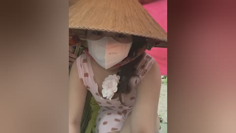 Media: Video of a Vietnamese woman in traditional attire, wearing a conical hat, white mask, and floral-patterned dress, kneeling in a room with pink walls, hands on the floor, looking forward.