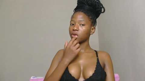 Video of a young Black woman with medium skin tone and braided hair in a high bun. She wears a black lace bra, revealing cleavage. She touches her lips with her left hand, looking contemplative against a plain beige background.