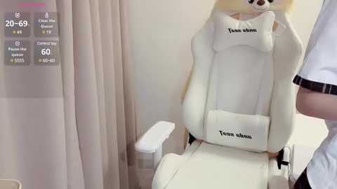 Media: Video of a white gaming chair with a stuffed dog head labeled \"Tonsu whin.\" Background includes beige curtains, a white armrest, and a man in a white shirt with black trim.