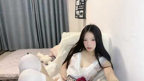 Media: A video of an East Asian woman with long black hair, wearing a white lace lingerie top, sitting on a bed with white sheets and plush toys.