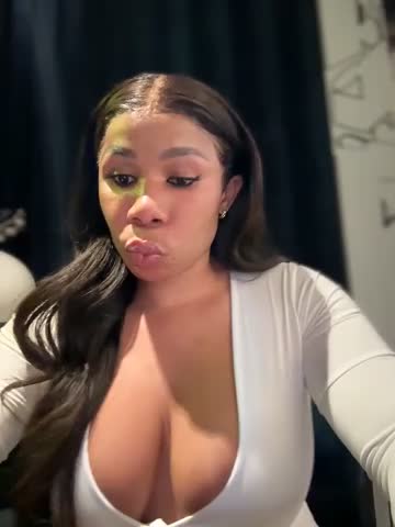 Media: A video of a curvy, light-skinned Black woman with long, wavy dark hair, wearing a low-cut white top that accentuates her large breasts. She's indoors, in front of a dark curtain, making a pouty face.