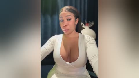 Media: Video of a Black woman with medium-brown skin, wearing a low-cut white top revealing ample cleavage, sitting indoors with a blurred background featuring a potted plant and a vase.