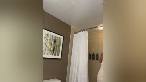 Media: Video of a modern bathroom featuring a beige wall, framed abstract art, a white shower curtain, and toiletries on a shelf, with a beige towel hanging nearby.