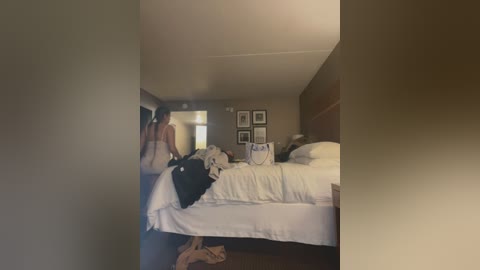 Media: A video of a dimly lit, neatly arranged hotel room featuring a king-sized bed with white linens, a brown headboard, and framed photos on the wall. A woman in a black top and jeans stands by the bed, holding a black bag.