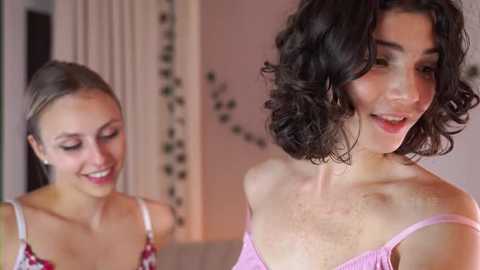 Media: Video of two smiling women, one with shoulder-length wavy brown hair, the other with straight brown hair, wearing a pink off-shoulder top. Soft lighting, cozy indoor setting with floral decor.