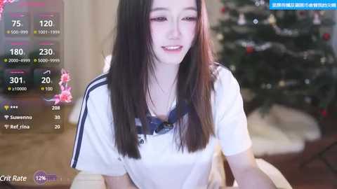 Media: A video of an East Asian woman with long black hair, fair skin, and a white T-shirt, smiling while sitting on a couch in a softly lit living room with a decorated Christmas tree in the background.