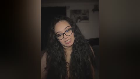 Media: Video of a smiling, light-skinned woman with long, curly black hair, wearing glasses, standing indoors in a dimly lit room with dark curtains.