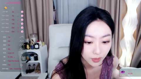 Media: Video of an East Asian woman with long black hair, light skin, and a pink blouse, sitting indoors. Background includes beige curtains, a white chair, and a cluttered shelf with bottles.