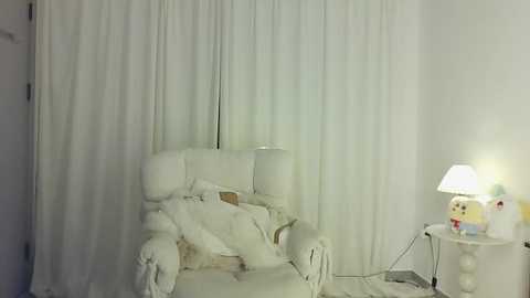 Media: A video of a white, plush armchair and matching ottoman in a minimalist room with white curtains and a small, white table lamp. The room exudes a serene, monochromatic atmosphere.