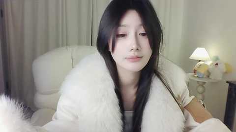 Media: A video of a young Asian woman with long black hair, fair skin, and a slender physique, wearing a white fur coat, sitting on a white leather chair in a softly lit room with beige curtains and a nightstand with a lamp and stuffed toys.