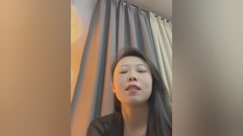 Media: A video of an Asian woman with long black hair, wearing a black top, standing in front of a gray and beige curtain, eyes closed, mouth slightly open, looking serene.