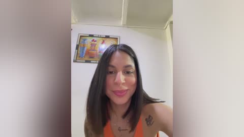 Media: Video of a Latina woman with straight, dark brown hair, wearing a bright orange tank top, smiling, with a small tattoo on her right shoulder, in a plain, white-walled room with a colorful poster on the wall.