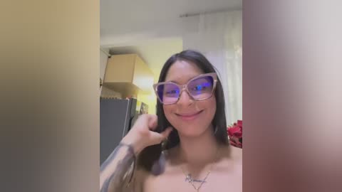 A video of a smiling, topless woman with long dark hair, wearing large glasses, standing in a kitchen with a beige wall and a white curtain.