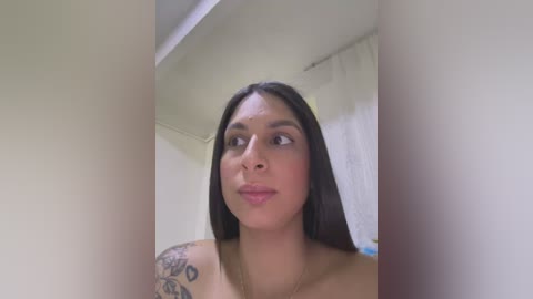 Media: Video of a Latina woman with long, dark hair and medium skin tone, wearing a gold necklace, looking upwards, showcasing a floral tattoo on her shoulder. She stands in a dimly lit bathroom with white walls and a shower curtain.