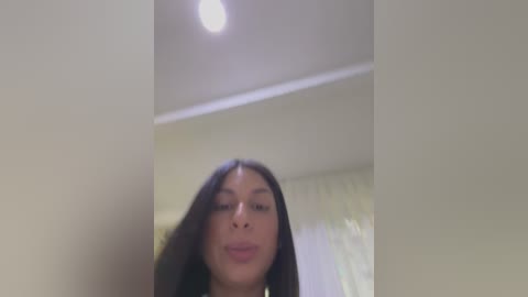 Media: A video of a young woman with long black hair, light brown skin, and a neutral facial expression, taken from a low angle, showing part of a ceiling light and a blurred background.