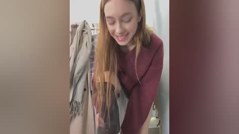 Media: Video of a young woman with long, straight, light brown hair, wearing a maroon sweater, bending over in a beige room with clothes hanging on a rack in the background.