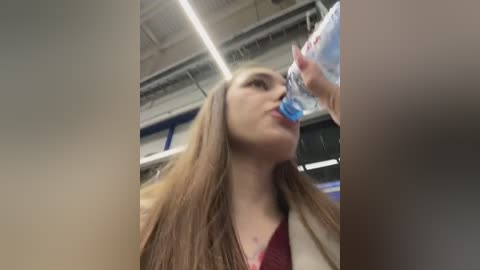 Media: A video of a young woman with long brown hair and light skin drinking from a blue water bottle in an industrial setting with fluorescent lights and exposed pipes in the background.