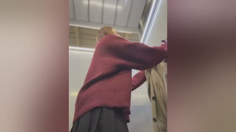 Media: A video of a blonde woman in a maroon sweater and black skirt, bending over to open a door. The background shows a beige wall and fluorescent lights.