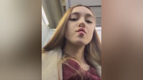 Media: Video of a young woman with straight, light brown hair, wearing a maroon sweater and beige coat, making a pouty face.