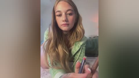 Media: Video of a young Caucasian woman with long blonde hair, wearing a light green jacket, applying lipstick in a dimly lit room.