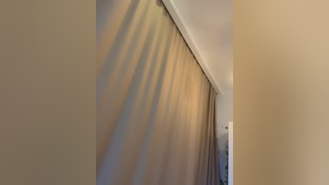 A video of a beige wall with a vertical shadow cast by a closed curtain, creating a striking diagonal pattern. The room's lighting is soft, with a hint of warm yellow light.