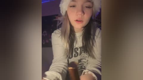 Media: A young woman with fair skin and long, straight hair, wearing a white Santa hat and a \"Budweiser\" t-shirt, holds a lit cigar in a dimly lit room.