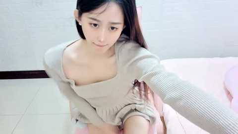 Media: Video of a young Asian woman with long black hair, fair skin, and a petite build, wearing a beige, off-shoulder knit sweater, sitting on a pink bed in a minimalist, brightly-lit room with white walls.