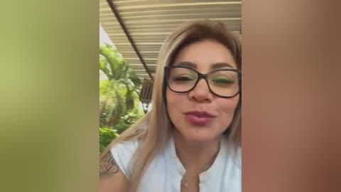 Media: A video of a smiling, light-skinned woman with long blonde hair, wearing black-rimmed glasses and a white blouse, taken outdoors under a beige canopy.