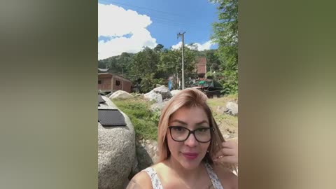 Media: A video of a smiling, light-skinned woman with shoulder-length, straight blonde hair and glasses, wearing a floral-patterned tank top, standing outdoors among rocks, trees, and a red-roofed building under a clear blue sky.