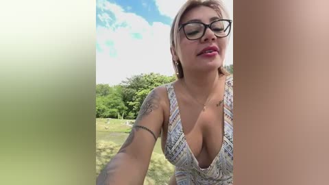 Media: Video of a fair-skinned woman with short blonde hair, wearing black-framed glasses and a low-cut, patterned top, outdoors with a lush green background and blue sky.