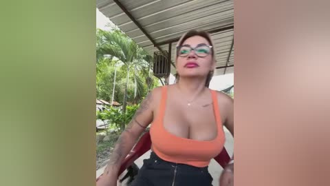 Media: Video of a curvy, middle-aged woman with large breasts, wearing a low-cut orange tank top and black high-waisted pants, standing outdoors under a patio roof with a green palm tree in the background.