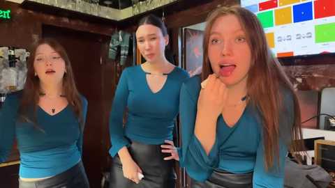 Media: Video of three Caucasian women in teal V-neck tops and black pants, posing in a dimly lit bar with a colorful digital sign and wooden walls.