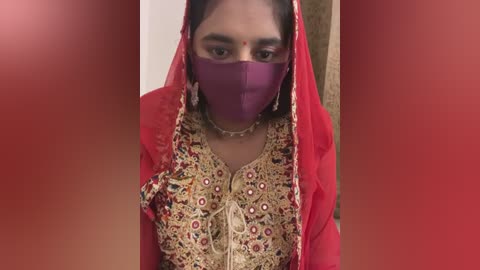 Media: Video of a South Asian woman with medium skin tone, wearing a red saree with intricate gold embroidery, a purple face mask, and a red dupatta.