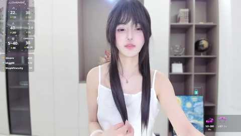 Media: A young East Asian woman with long black hair, pale skin, and a slim physique, wearing a white spaghetti-strap top, stands in a modern, minimalist room with a wall-mounted clock and a bookshelf in the background.