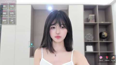 Media: A video of an East Asian woman with straight black hair, fair skin, and wearing a white spaghetti-strap top, standing in a modern, minimalist room with grey shelves and a globe.